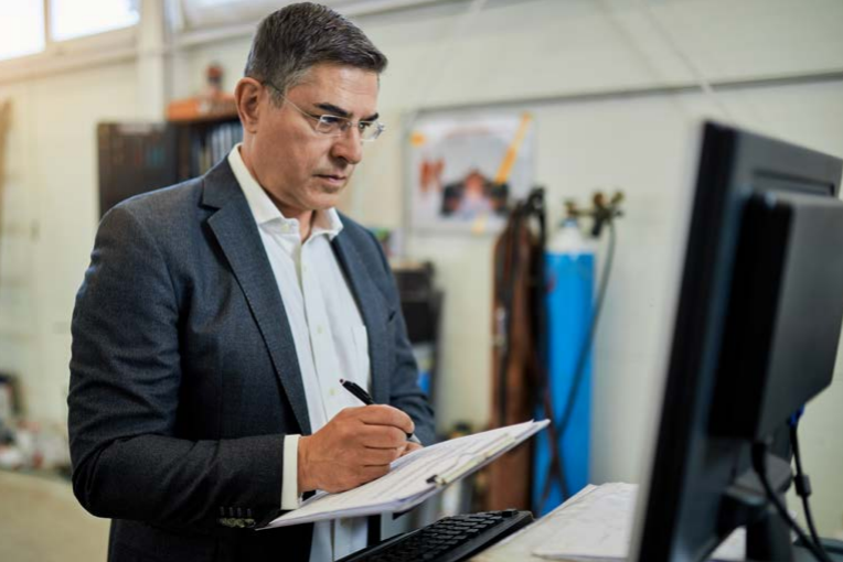 Man writing on clipboard looking at computer. 